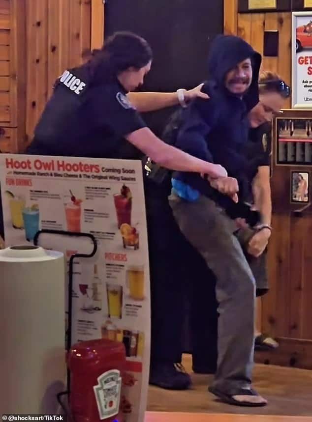 Violent altercation between customers, police officer intervening at a bar or restaurant, tempers flare, conflict, security situation, law enforcement, disorderly conduct, humorous or chaotic scene, internal location with wood paneling, promotional stand for Hoot Owl Hooters drinks, casual attire, tense moment.